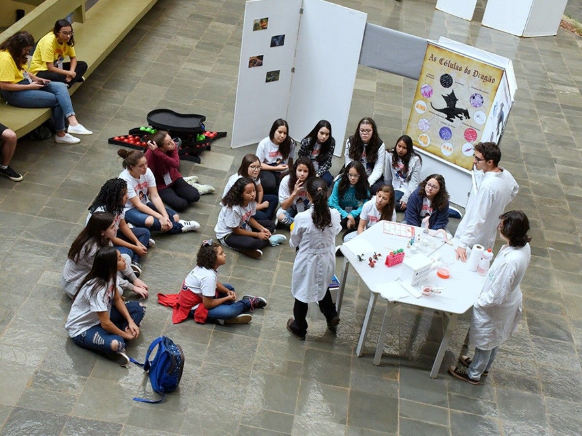 A foto mostra um grupo de alunas no entorno de uma cientista que conversa com elas