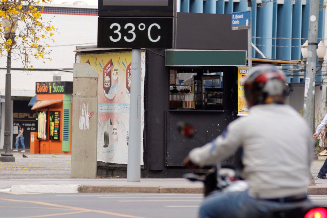 A imagem mostra um motociclista, de capacete vermelho e preto, jaqueta bege claro e calça jeans, passando em frente a um termômetro eletrônico que marca 33ºC.
