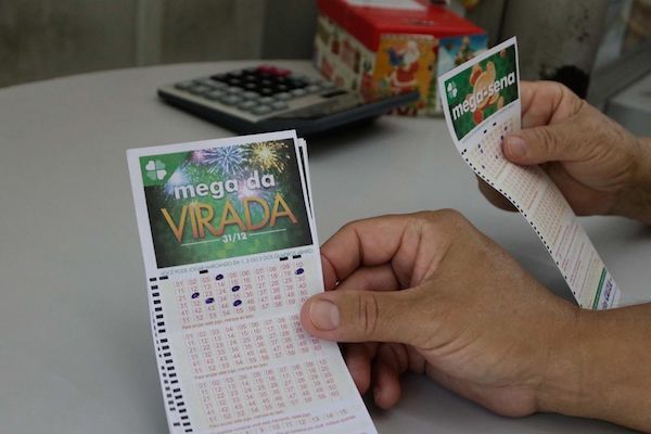 A foto mostra uma mão segurando um volante da Mega da Virada dentro de uma lotérica.