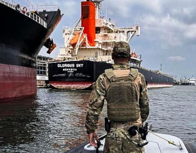 Na foto é possível ver um militar, vestido com um uniforme camuflado, em uma pequena embarcação no porto de Santos. Ao lado, há a lateral proa de um navio. A frente, há outra embarcação.de grande porte.