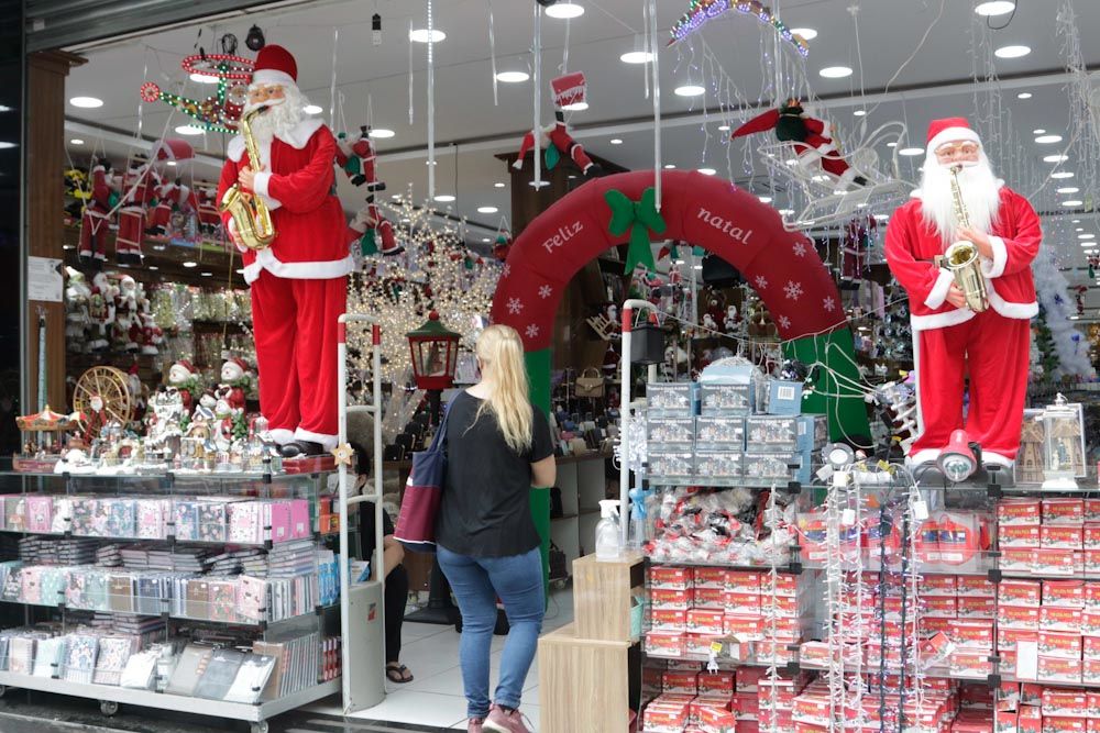A foto mostra a fachada de uma loja repleta de enfeites natalinos e com dois bonecos de Papai Noel tocando saxofone. Uma mulher loira, de blusa preta e calça jeans, entra no comércio segurando uma bolsa.