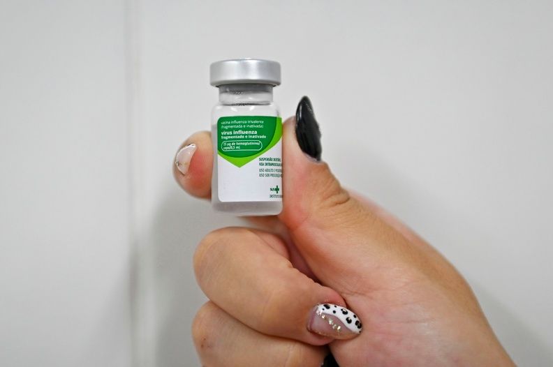 A foto mostra uma mão com unhas longas, com detalhes em preto e branco, segurando uma ampola de vacina contra a gripe. O vidro é transparente, com uma tampa metalizada e um adesivo verde e branco