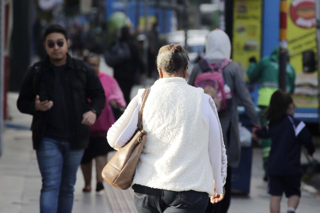 a foto mostra várias pessoas andando em uma calçada, vestindo blusas de frio