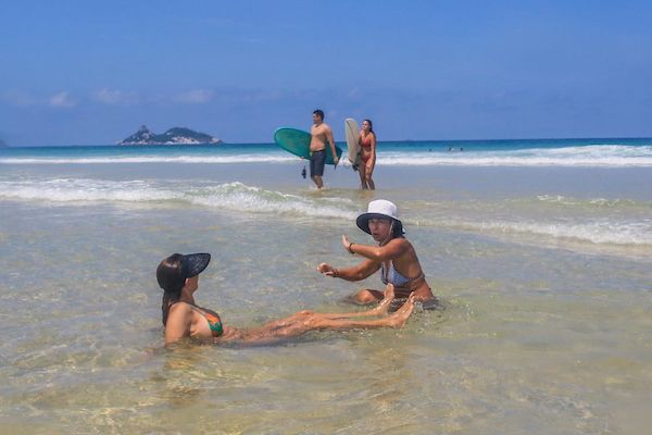 A foto mostra duas mulheres sentadas na beira do mar e no fundo duas pessoas caminhandos na areia. Uma delas segura uma prancha