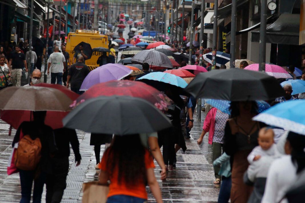 a foto mostra movimentação de pessoas segurando guarda-chuvas em um calçadão no centro