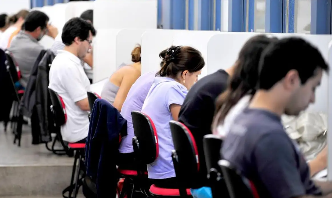 a foto mostra várias pessoas prestando prova de um concurso público em guichês