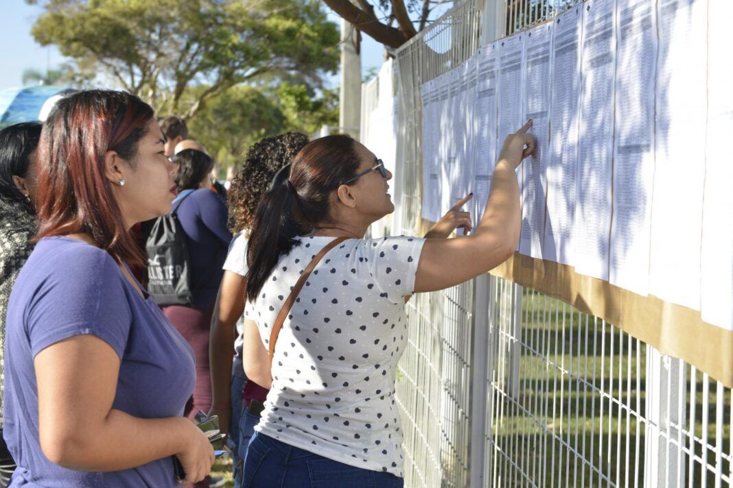 A foto mostra pessoas olhando uma lista anexada em um portão de local de prova