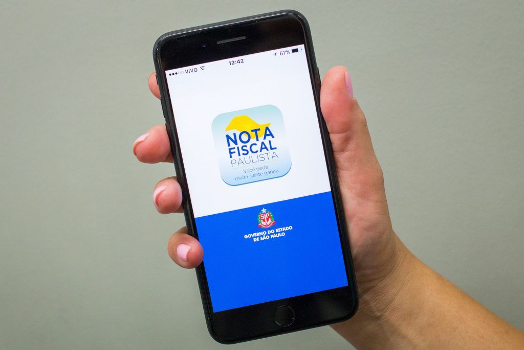 A foto mostra uma mão segurando um celular que esta com o aplicativo da Nota Fiscal Paulista na tela