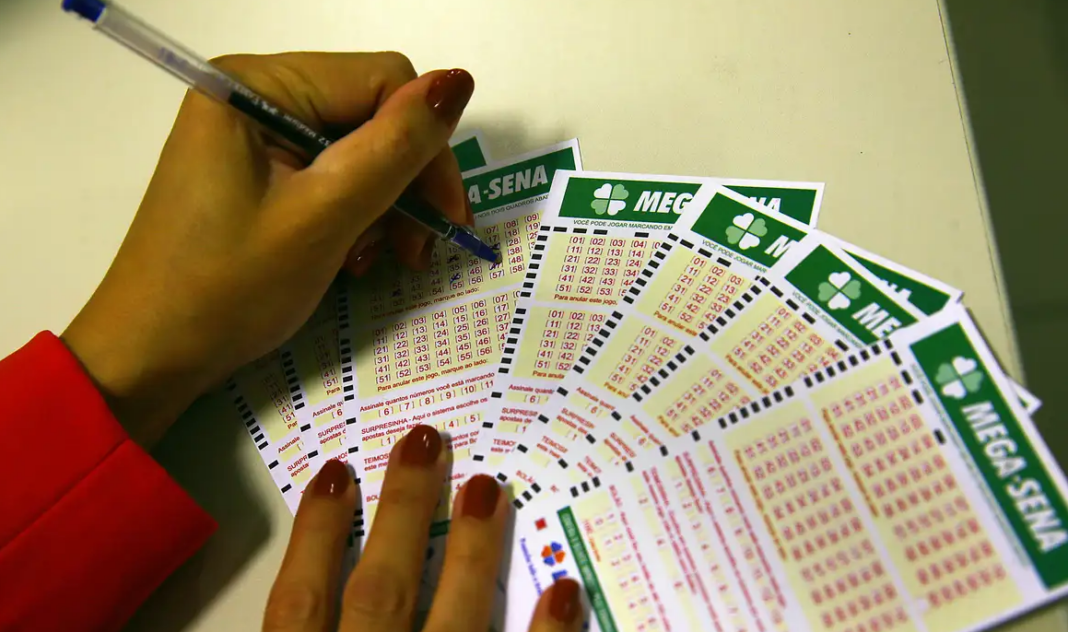 a foto mostra a mão de uma pessoa segurando uma caneta e fazendo marcações em cartões de aposta da Mega-Sena