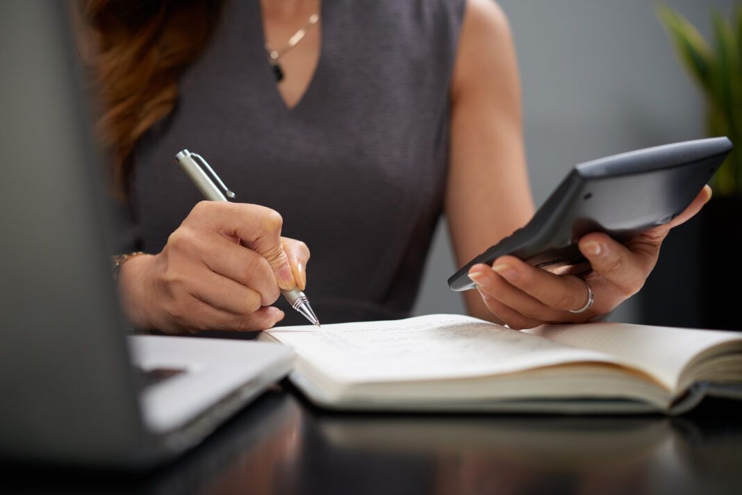 a foto mostra parte do corpo de uma mulher com uma calculadora na mão e escrevendo em um papel