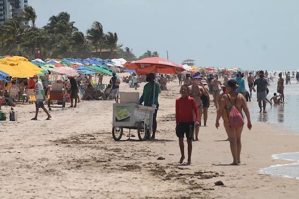 A foto mostra pessoas na praia, em guarda-sol e também caminhando na faixa de areia, além de carrinho de vendedor ambulante