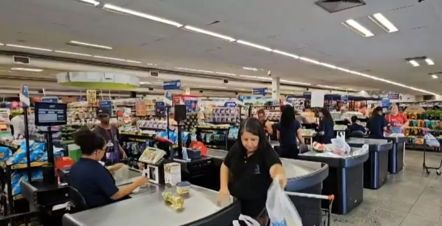 A foto mostra pessoas circulando e passando pela área de caixas de um supermercado