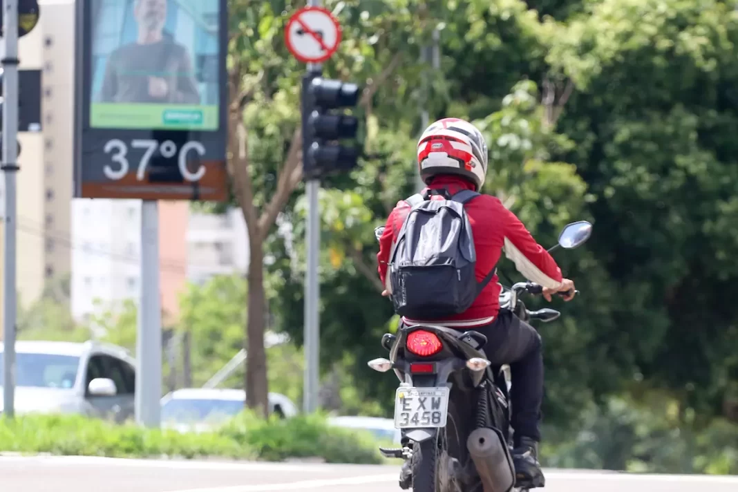 A foto mostra um motociclista passando por uma via e um termometro marcando 37ºC ao fundo