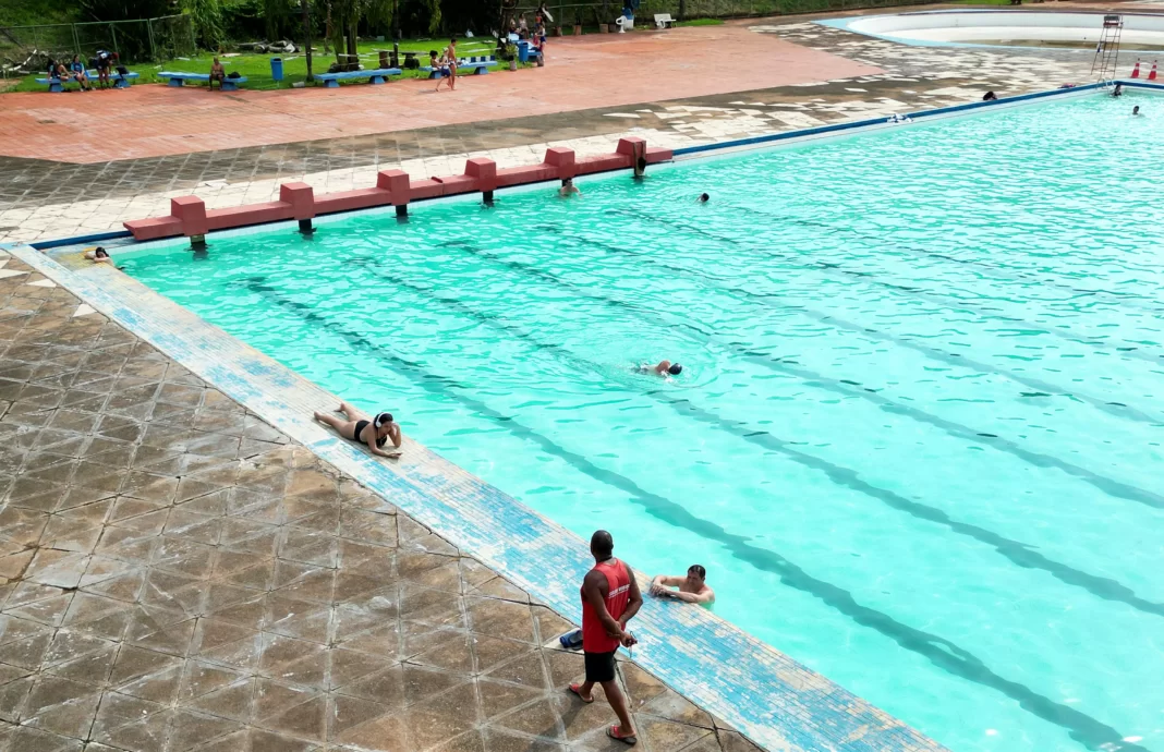 a foto mostra uma piscina com pessoas dentro e um salva-vidas caminhando fora