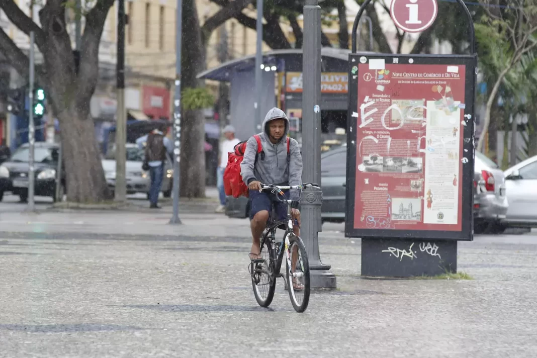a foto mostra uma pessoa andando de bicicleta passando por uma praça da cidade no meio de chuva