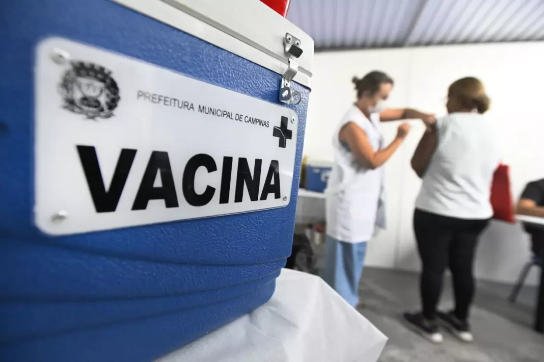 a foto mostra uma caixa térmica em primeiro plano com vacinas e atras uma pessoa sendo vacinada