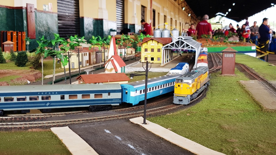 Maquete ferroviária reproduz uma cidade. No primeiro plano estão vagões de passageiros azuis-claros e uma locomotiva amarela. Ao fundo, prédio de estação, galpões, árvores e paisagem.