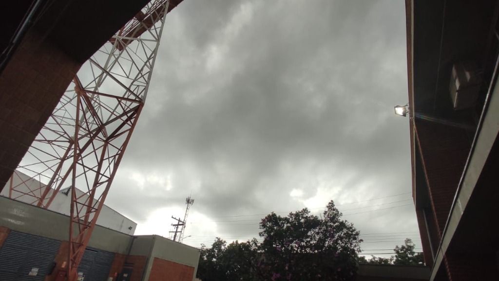 A foto mostra um céu nublado e cinza, com árvore e uma torre de transmissão ao fundo