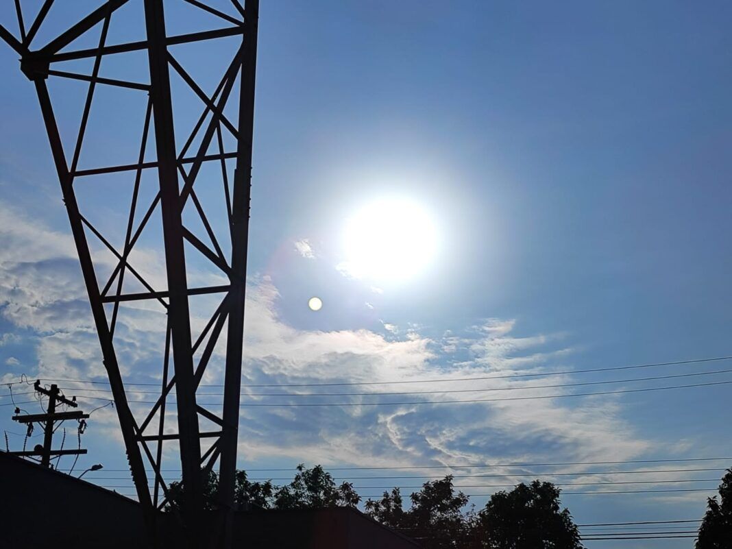 A foto mostra o sol brilhando forte em um céu azul, com algumas nuvens brancas. Há sombra de uma árvore e uma torre de transmissão