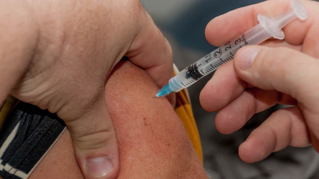 Mãos sem luvas injetam vacina no topo do braço de uma pessoa de cor branca.