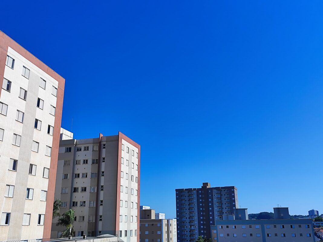 A foto mostra fachadas de prédios e um céu azul, sem nuvens e com sol
