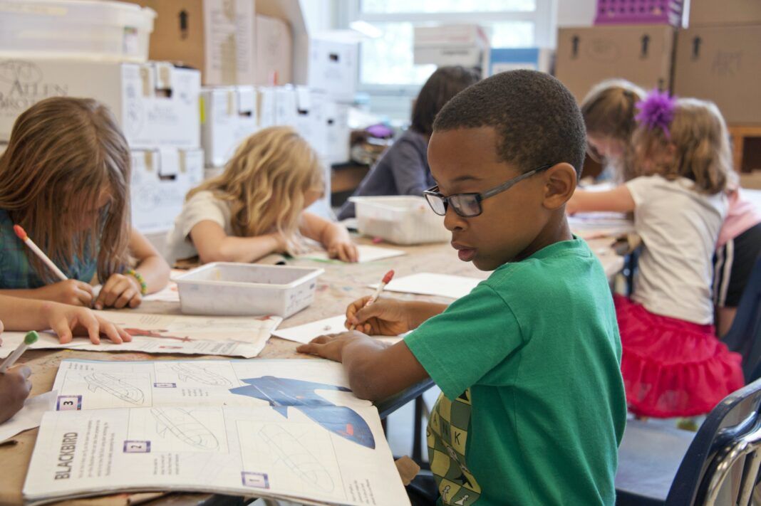 A foto mostra uma criança negra escrevendo e outras crianças ao redor