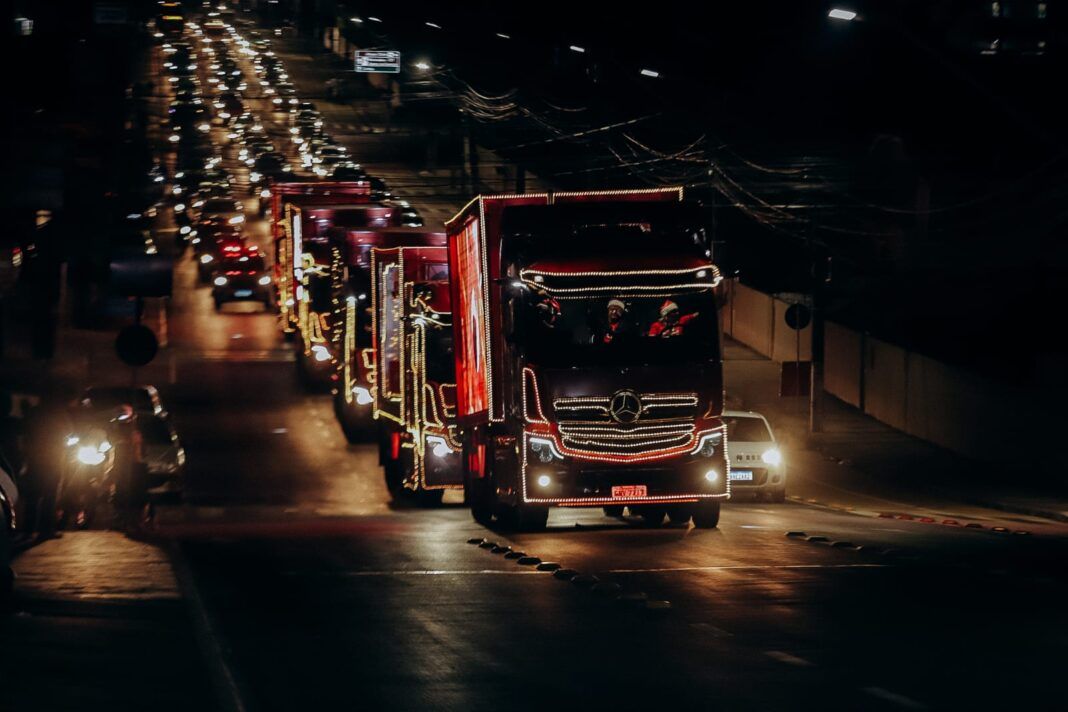 A foto mostra caminhões iluminados com luzes natalinas e uma multidão de pessoas