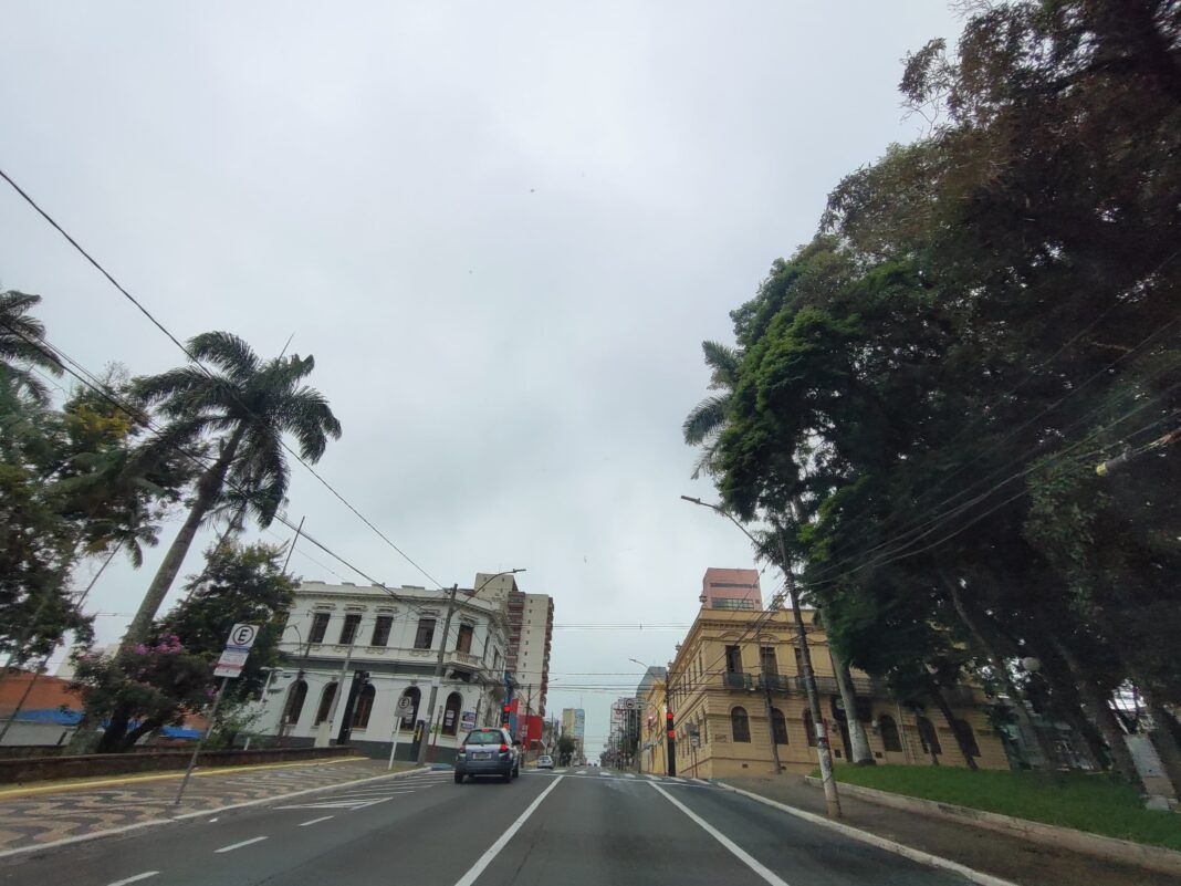 A foto mostra uma avenida com o tempo nublado, duas árvores e dois prédios históricos, um branco e outro amarelo