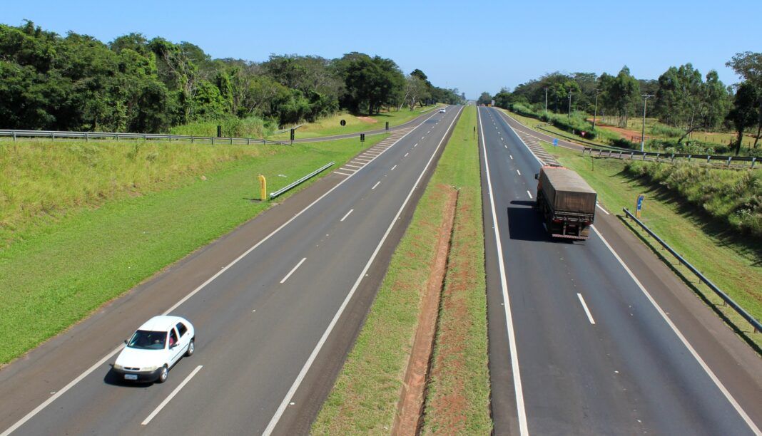 A foto mostra uma rodovia com um caminhão e ummcarro passando