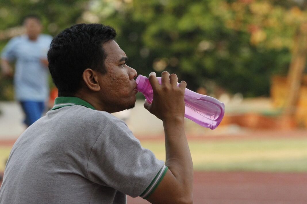 A foto mostra um homem moreno bebendo água em uma garrafa cor de rosa