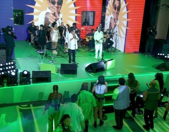 Palco verde do Big Brother Brasil com Zeca Pagodinho e Pretinho da Serrinha se apresentando, com os participantes na frente do palco