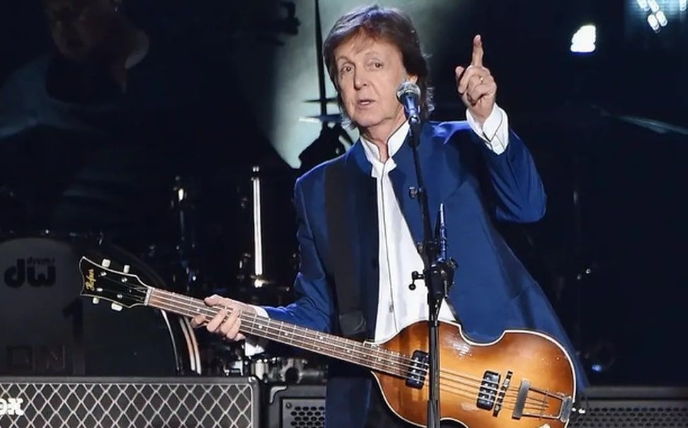 A foto mostra o cantor Paul McCartney no palco com blazer azul e camisa branca e com uma guitarra